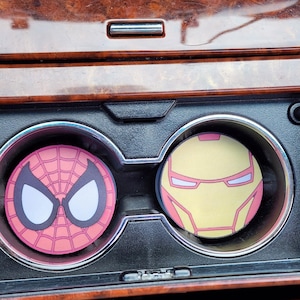 Puede incluir: Dos posavasos para coche con diseños de superhéroes. Uno muestra la cara de Spider-Man en rojo y blanco, el otro la máscara de Iron Man en amarillo y rosa. Los posavasos están en los portavasos de un coche.