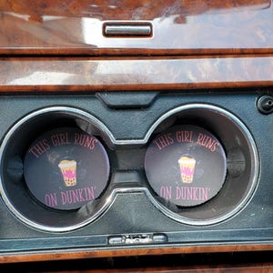 Könnte beinhalten: Zwei runde Auto-Untersetzer mit dem Text "THIS GIRL RUNS ON DUNKIN'" in Rosa. Jeder Untersetzer zeigt eine Grafik eines Bubble Tea-Getränks. Die Untersetzer befinden sich in einem Getränkehalter im Auto.