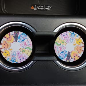 Op de afbeelding: Twee ronde onderzetters voor in de auto met een kleurrijk cartoonontwerp van verschillende wezens. De onderzetters zijn geplaatst in de bekerhouders van de auto, met een zwart dashboard en zilveren afwerking.