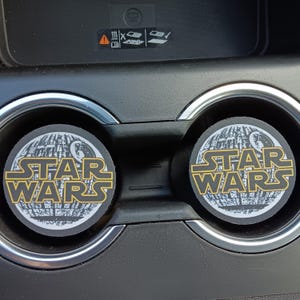 May include: Two black car coasters with the words "STAR WARS" in yellow and white lettering. The coasters have a circular design with a planet graphic in the background. The coasters are in a car cup holder.