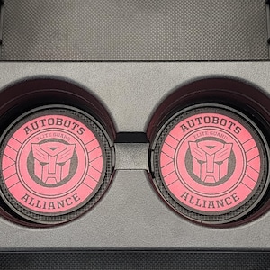 May include: Two pink car coasters with the Autobots logo, featuring a shield design and the words "AUTOBOTS ELITE GUARD ALLIANCE". The coasters are circular and sit in a car's cup holders.