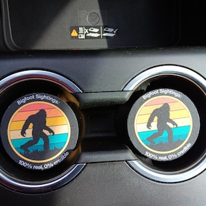 Puede incluir: Dos posavasos redondos para coche con una silueta de Bigfoot en un diseño retro de puesta de sol. Los posavasos dicen "Bigfoot Sightings" y "100% real, 0% credible."