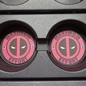 May include: Two car cup holder coasters with a Deadpool design. The coasters are circular with a red border and the words "MAXIMUM EFFORT" around the edge. The center features the Deadpool logo in black and white.