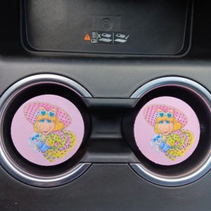 Peut inclure: Deux dessous de verre roses pour voiture avec un personnage de dessin animé portant un grand chapeau rose à pois et des lunettes de soleil en forme de cœur. Le personnage tient un petit animal en peluche bleu et porte une robe à pois jaune et verte.