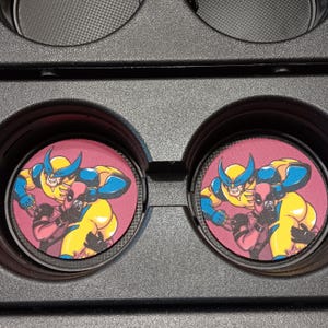 May include: Set of two car coasters featuring a comic-style illustration of Wolverine and Deadpool embracing. The coasters have a maroon background and are set in a black car console.
