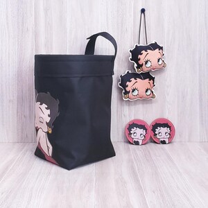 May include: Black tote bag, two hanging ornaments, and two coasters featuring a cartoon character. The tote bag has a handle and a character graphic on the front. The ornaments and coasters have the same character design.