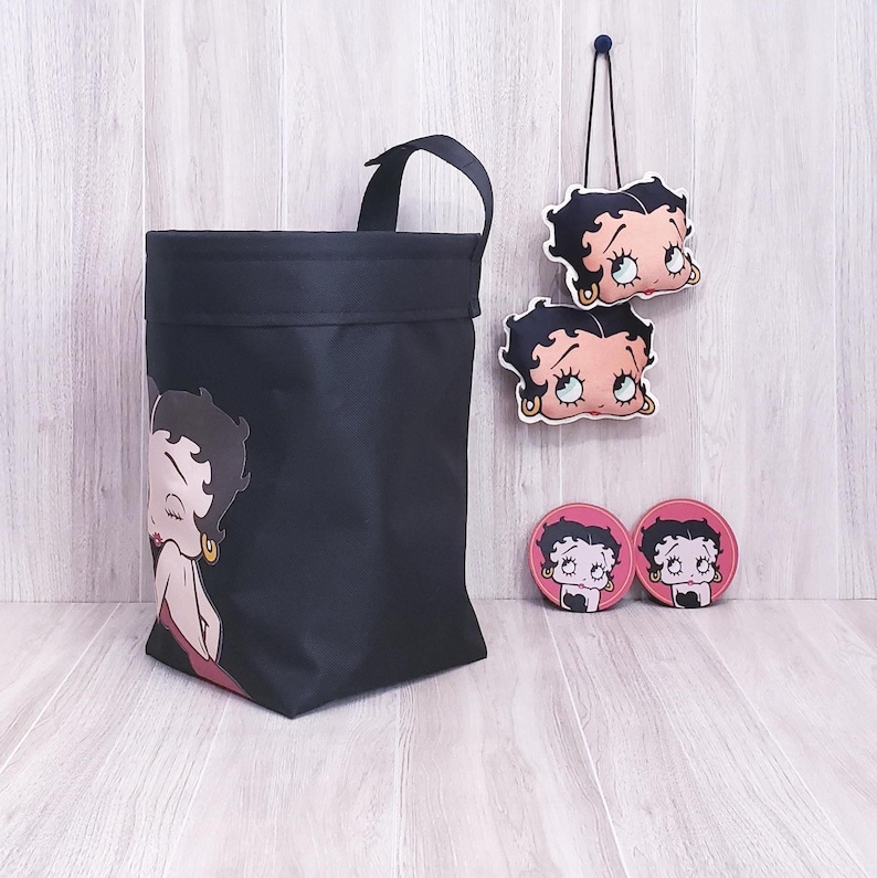 May include: Black tote bag, two hanging ornaments, and two coasters featuring a cartoon character. The tote bag has a handle and a character graphic on the front. The ornaments and coasters have the same character design.