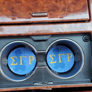 May include: Two blue car coasters with the Greek letters Sigma, Gamma, and Rho in yellow. The coasters are in a car's cup holders, set against a wood-grain dashboard.