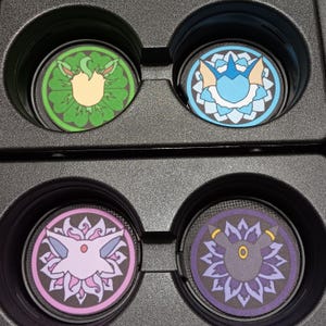 May include: Four round car coasters with colorful designs. The coasters feature various cartoon character designs in green, blue, pink, and purple. The coasters are set in a black car console.
