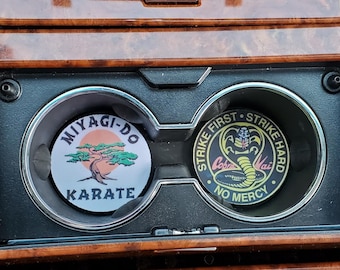 Puede incluir: Dos posavasos para coche con temas de Karate Kid. Un posavasos tiene el logo de Miyagi-Do Karate con un árbol y el texto "KARATE". El otro posavasos tiene el logo de Cobra Kai con una serpiente y el texto "STRIKE FIRST, STRIKE HARD, NO MERCY".