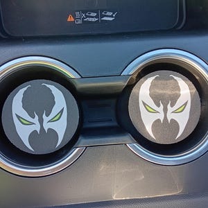 Könnte beinhalten: Zwei graue Auto-Untersetzer mit dem Logo der Comicfigur Spawn. Das Logo zeigt ein weißes Gesicht mit grünen Augen auf schwarzem Hintergrund. Die Untersetzer befinden sich in den Getränkehaltern eines Autos.