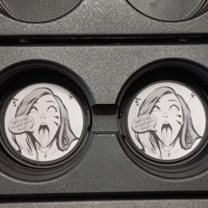 以下が含まれることがあります： 黒いトレイには、舌を出した女性の漫画イラストが描かれた2つの円形容器が入っています。「Spill it all! I'll catch every drop!」というテキストが吹き出しの中にあります。