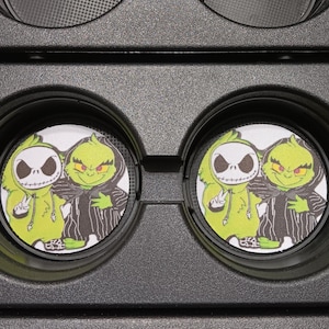 May include: Set of four car coasters featuring a cartoon image of Jack Skellington and the Grinch. The coasters are round, with a white background and a black border. The Grinch is wearing a black and white striped robe and Jack is wearing a green hoodie.