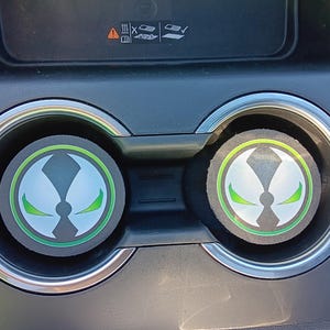 May include: Two car coasters with the Spawn logo in white and green on a black background. The coasters are circular and fit into the car's cup holders. The design is a stylized face with green eyes and a green outline.