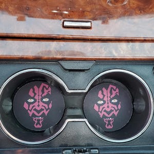 Puede incluir: Dos posavasos negros para coche con un gráfico magenta y blanco de la cara de Darth Maul. Los posavasos son circulares y encajan en los portavasos del coche. El interior del coche tiene un acabado de vetas de madera.