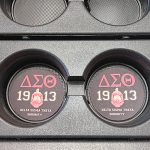 May include: Two black car cup holder coasters with a Delta Sigma Theta Sorority design. The coasters are black with a pink and white emblem, the Greek letters, and the year 1913. The text "DELTA SIGMA THETA SORORITY" is printed on the bottom.