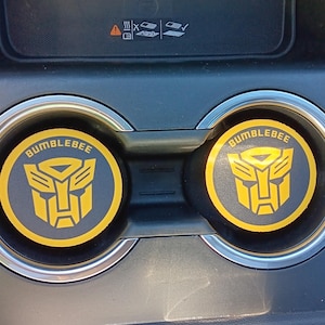 May include: Two yellow and black Bumblebee car coasters. Each coaster features the Bumblebee logo and the word "BUMBLEBEE" in yellow on a black background. The coasters are in a car's cup holders.