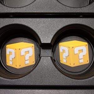 Puede incluir: Dos posavasos negros para coche con un diseño de caja amarilla con signo de interrogación. Los posavasos redondos encajan en los portavasos del coche. El diseño es de un videojuego.