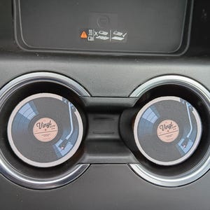 May include: Two round car coasters with a vinyl record design. The coasters are black with a light pink label that reads "Vinyl". The design includes a turntable illustration. The coasters are placed in a car's cup holders.