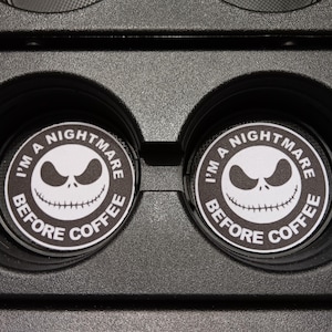 May include: Two black and white car coasters with the text "I'M A NIGHTMARE BEFORE COFFEE" around a Jack Skellington face. The coasters are circular and fit into a car's cup holders.