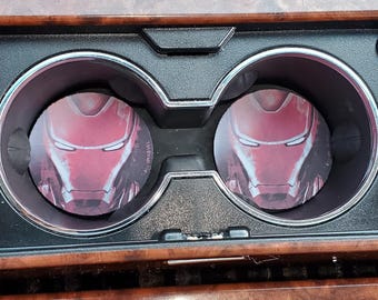 May include: Two round car coasters featuring the Iron Man helmet design, placed in a car's cup holders. The coasters are red and gray, with a detailed graphic of the superhero's face. The car's interior has a wood grain finish.