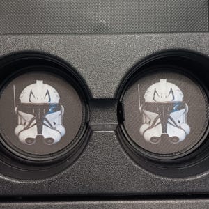 May include: Two black car coasters featuring a white and black illustration of a Clone Trooper helmet. The coasters are circular and fit into a car's cup holders. The design is a popular reference from a science fiction franchise.