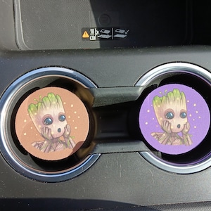 以下が含まれることがあります： 木のような外観の漫画のキャラクターが特徴の車のコースター2枚。1つのコースターは白いドットが付いた茶色で、もう1つは紫です。キャラクターは大きな青い目を持ち、手で顔を支えています。