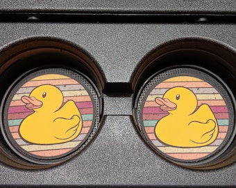 May include: Two car coasters with a yellow rubber duck design. The duck is centered on a background of horizontal stripes in shades of orange, pink, and green. The coasters are set in a black car console.