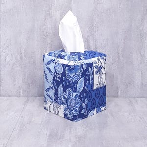 May include: A square tissue box cover with a blue and white floral pattern. The fabric design features various floral and leaf motifs in shades of blue on a white background. White tissues are visible extending from the top opening.