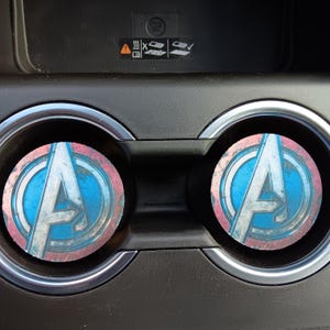 Puede incluir: Dos posavasos redondos para coche con el logotipo de los Vengadores en azul, blanco y rojo. Los posavasos están colocados en los portavasos de un coche, con un interior negro y adornos plateados.