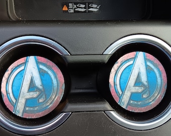May include: Two round car coasters featuring the Avengers logo in blue, white, and red. The coasters are placed in a car's cup holders, with a black interior and silver trim.