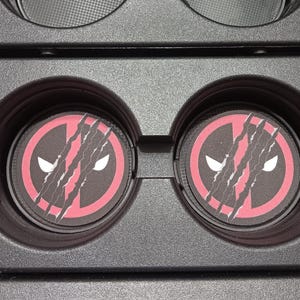 May include: Two black car coasters with a red and black Deadpool logo. The logo features a stylized face with white eyes and claw marks. The coasters are in a black car console.