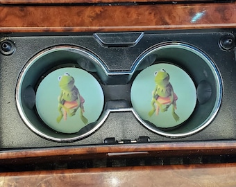May include: Two round car coasters featuring Kermit the Frog. The coasters are light green with a cartoon image of Kermit wearing a pink shirt. The coasters are placed in the car's cup holders, which are surrounded by a wood-grain finish.