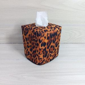 May include: A square tissue box cover with a brown and black leopard print pattern. White tissues are visible at the top. The cover is made of fabric and sits on a light wood surface.