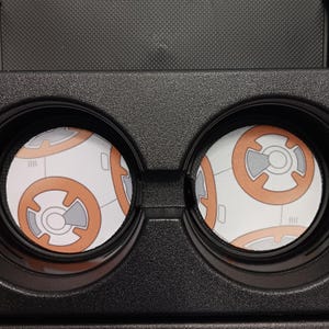 May include: Black VR headset with two circular lenses displaying a BB-8 droid design. The BB-8 design features orange and gray details on a white background. The headset is made of black plastic.
