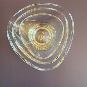 May include: Clear glass bowl set with a unique triangular shape. The set includes three nested bowls, each with a rounded triangular design. The bowls have a smooth, transparent appearance, and the bottom bowl has etched markings.