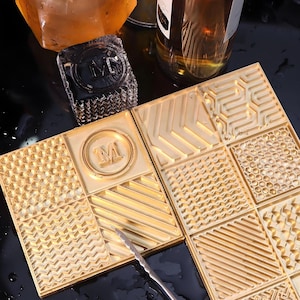 Può includere: Due stampi per ghiaccio rettangolari color oro con varie texture geometriche, tra cui motivi a nido d'ape, linee diagonali e motivi a chevron. Uno stampo presenta un emblema circolare con la lettera M al centro. È visibile una pinza in metallo.