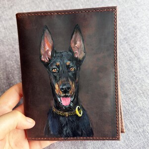 May include: A brown leather wallet with a painted Doberman Pinscher portrait. The dog has black fur, brown eyes, and a pink tongue. The wallet is held by a hand and has a stitched border. The wallet is approximately 10 cm in height.