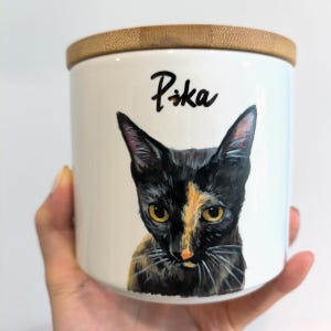 May include: A white ceramic jar with a bamboo lid. The jar features a hand-painted portrait of a cat with black and orange fur and yellow eyes. The name "Pika" is written above the cat.