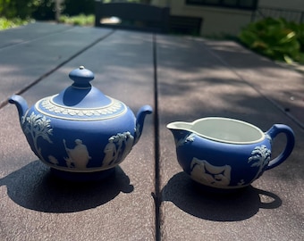 May include: A blue and white jasperware sugar bowl with a lid and a matching creamer. Both pieces feature matte blue surfaces with white relief neoclassical figures and botanical motifs. The sugar bowl has a round lid with a small knob handle.