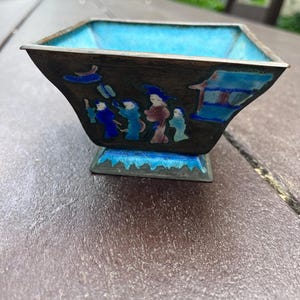 May include: A square bowl with a turquoise interior and a dark exterior. The bowl features a painted scene of figures and has a silver rim and a decorative base. The design showcases a blend of colours and intricate details.
