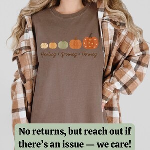 Puede incluir: Camiseta marr&oacute;n con una fila de cinco calabazas de varios colores y dise&ntilde;os. El texto "Healing, Growing, Thriving" est&aacute; impreso debajo. La camiseta se lleva con una camisa a cuadros.