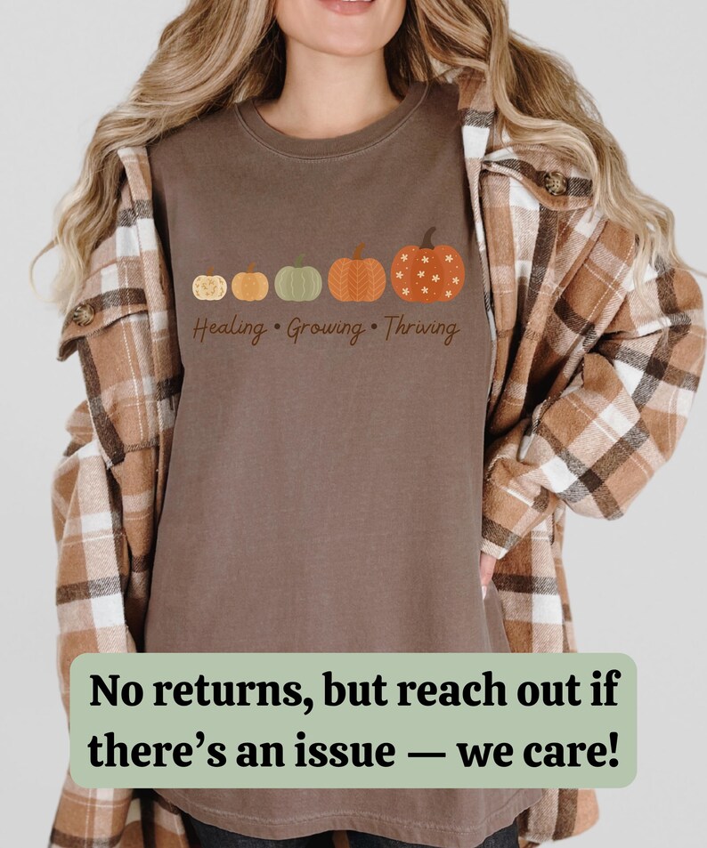 Puede incluir: Camiseta marr&oacute;n con una fila de cinco calabazas de varios colores y dise&ntilde;os. El texto "Healing, Growing, Thriving" est&aacute; impreso debajo. La camiseta se lleva con una camisa a cuadros.