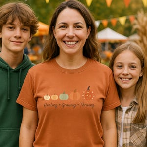 Puede incluir: Una camiseta naranja con un dise&ntilde;o de calabaza y las palabras "Healing, Growing, Thriving". La camiseta la lleva una persona de pie con otras dos personas. El fondo incluye banderas de banderines de colores y follaje oto&ntilde;al.