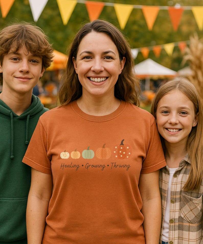 Puede incluir: Una camiseta naranja con un dise&ntilde;o de calabaza y las palabras "Healing, Growing, Thriving". La camiseta la lleva una persona de pie con otras dos personas. El fondo incluye banderas de banderines de colores y follaje oto&ntilde;al.