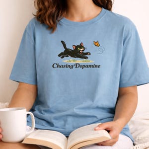 May include: Light blue t-shirt featuring a black cat chasing a butterfly. The cat has green eyes and a red collar. The text "Chasing Dopamine" is printed below the cat illustration. The shirt is a solid color and has short sleeves.