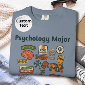 May include: A light blue t-shirt with the words "Psychology Major" and various psychology-related graphics. The graphics include a brain, a mouse, and the words "Therapy" and "Mental Health". A white circle with the words "Custom Text" is on the top left.