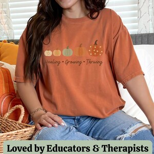 Puede incluir: Camiseta naranja quemado con un dise&ntilde;o de calabaza y las palabras "Healing, Growing, Thriving". El dise&ntilde;o incluye cuatro calabazas. La camiseta se usa con vaqueros azules.