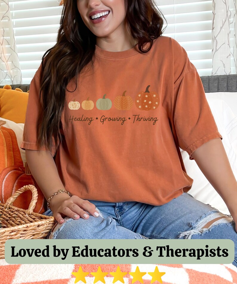 Puede incluir: Camiseta naranja quemado con un dise&ntilde;o de calabaza y las palabras "Healing, Growing, Thriving". El dise&ntilde;o incluye cuatro calabazas. La camiseta se usa con vaqueros azules.