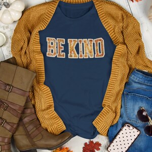 May include: A navy blue Comfort Colors tee with the words "BE KIND" in a floral design. The tee is paired with a mustard yellow cardigan, brown boots, and blue jeans. A phone with a polka dot case and sunglasses are also in the image.